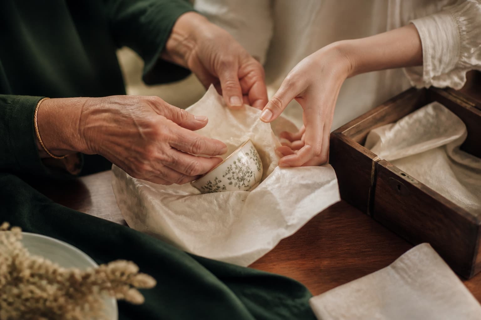 Two generations of hands wrapping a family teacup in tissue paper at a wooden table.